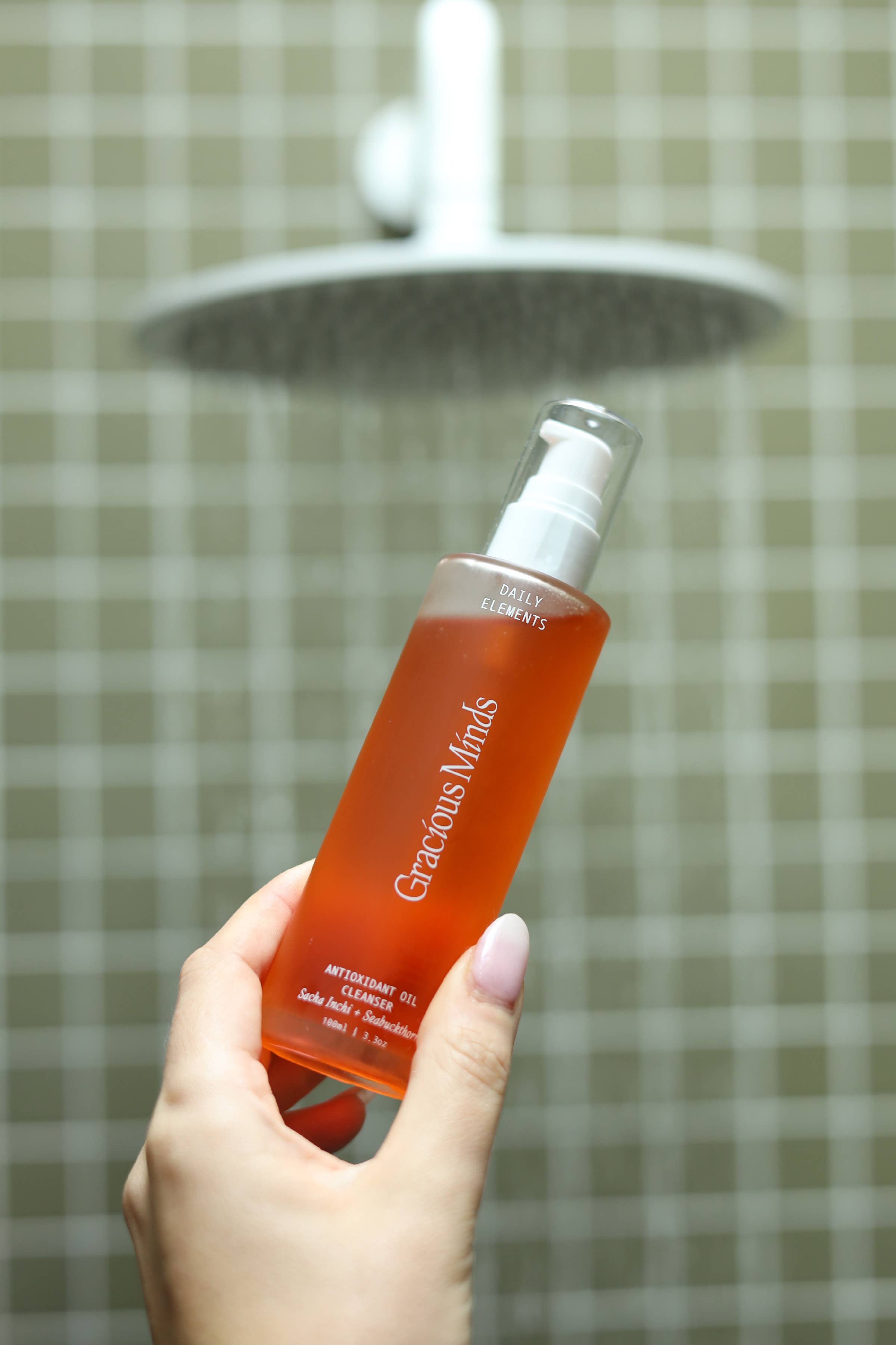 Gracious Minds Antioxidant oil cleanser orange liquid in bottle in front of green tiles held in a person hand